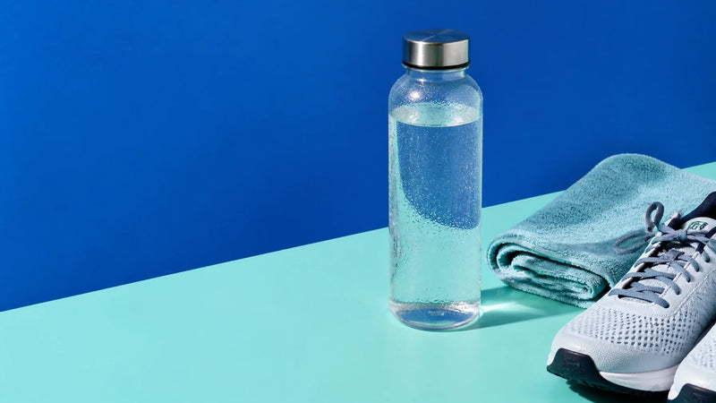 Clear reusable water bottle beside a gym towel and running shoes on a bold blue and aqua background